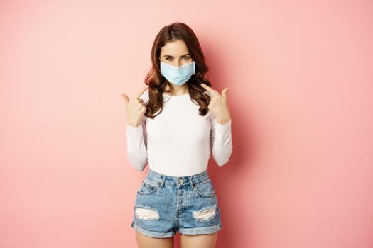 Portrait of young stylish woman wearing and pointing fingers at her medical face mask, smiling, preventive measures during covid 19 pandemic, quarantine, standing over pink background.