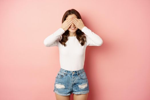 Cute happy girl waiting for surprise with closed eyes, blindfolded anticipating presents on valentines day, pink background.