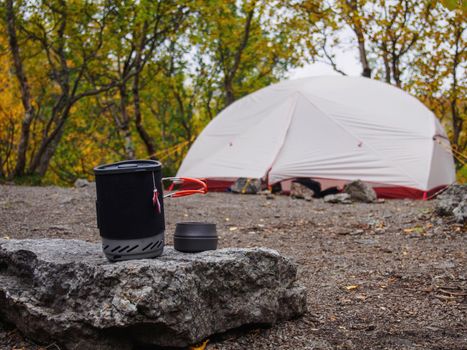 A pot for a gas burner with a mug sits on a stone against the background of a tent