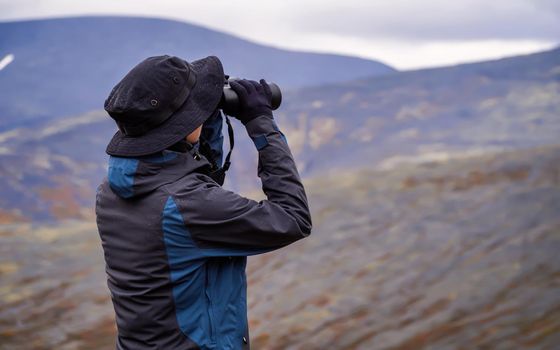 A woman in a hat on a mountain slope looks through binoculars in autumn against the backdrop of a lake and mountains. view from the back.