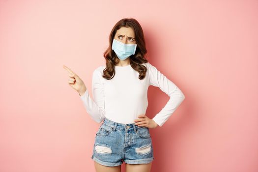 Confused, indecisive young woman in medical face mask, pointing finger left and looking complicated, making decision, standing over pink background.