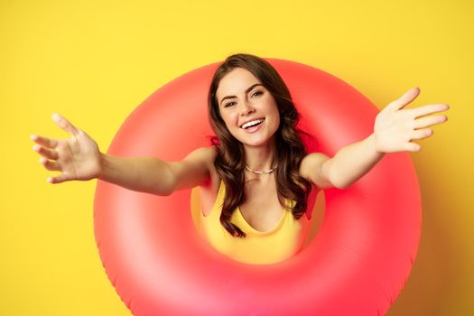 Close up portrait of happy young woman inside pink swimming ring, reaching hands forward, inviting go to the beach, enjoy holiday, standing over yellow background.