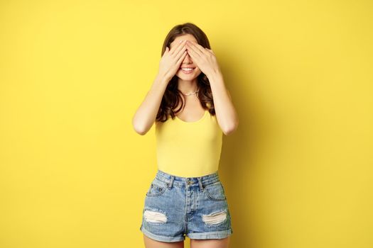 Portrait of brunette woman cover eyes with hands, blindfolded, smiling with anticipation, expecting smth, waiting for surprise, standing over yellow background.