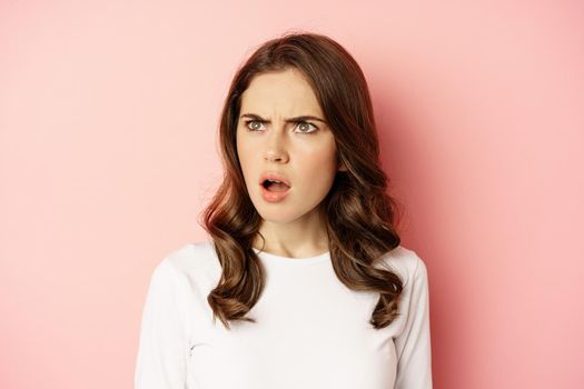 Close up portrait of confused, frustrated young woman, looking left with disappointment, puzzled by something, standing over pink background.