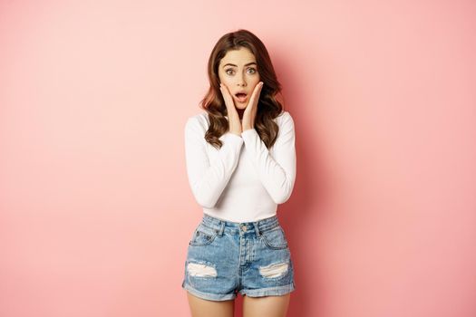 Shocked glamour girl looking at camera, gasping amazed. Woman staring surprised at advertisement, store discounts, standing over pink background.