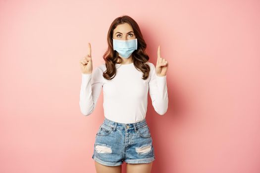 Portrait of young glamour girl in medical face mask, reading promo text, pointing and looking up, standing over pink background. Covid and pandemic concept