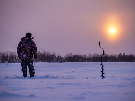 A young man fishes from a hole in the ice at dawn. winter fishing