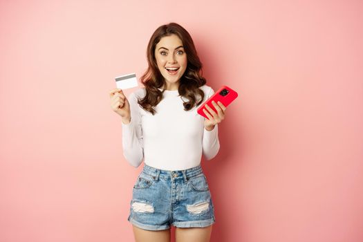 Enthusiastic brunette girl showing credit card and using mobile phone to order or pay, online shopping app, standing over pink background.