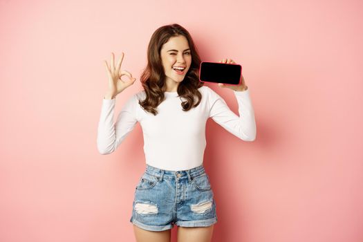 Happy winking girl demonstrating company store, mobile app, showing smartphone horizontally and okay, good sign, smiling happy, standing over pink background.