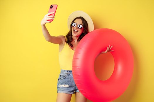 Stylish brunette girl on vacation, taking selfie with swim ring, going on beach, swimming in sea on summer holiday, standing over yellow background.