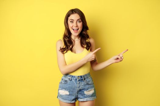Beautiful young woman pointing fingers at banner, logo or advertisement, smiling amazed, standing in summer clothes, yellow background.