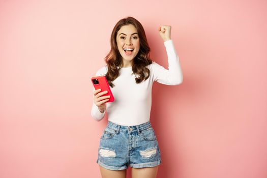 Enthusiastic brunette girl winning on mobile phone, holding smartphone and rejoicing, scream in joy, achieve app goal, pink background.