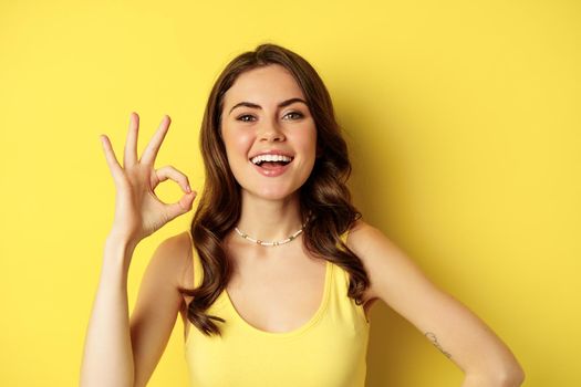 Close up portrait of young happy pretty woman, laughing and smiling with white teeth, showing excellent, okay ok sign, praise and compliment smth, yellow background.