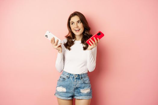 Technology, spring promo concept. Stylish glamour woman holding two smartphones, mobile phones in both hands, smiling pleased, buying new cellphone, pink background.