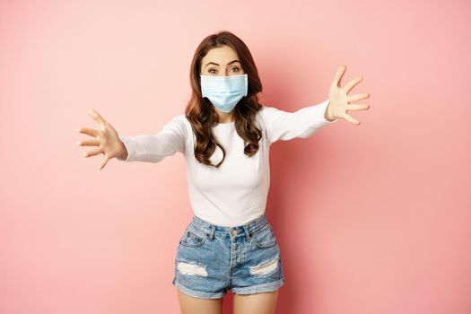 Portrait of smiling friendly girl in medical face mask, reaching hands, stretching arms forward for hug, receiving smth, standing over pink background.