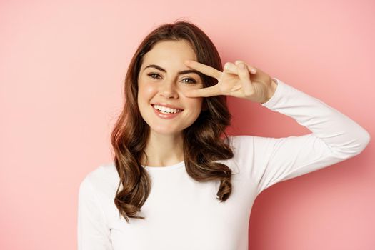 Close up portrait of coquettish young woman smiling, showing peace, v-sign gesture and posing happy, standing against pink background.