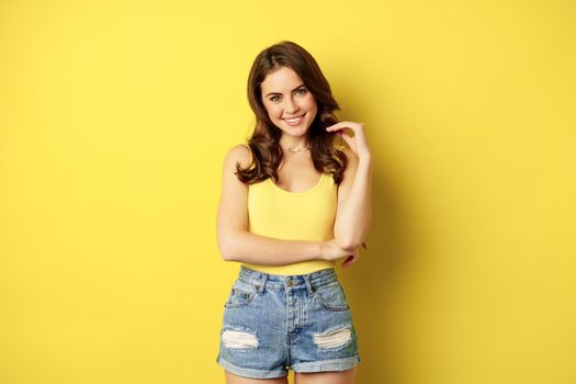 Summer holidays and people. Stylish bright girl in tank top and shorts, posing against yellow background, smiling and looking relaxed.