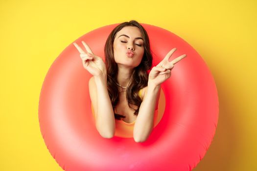Portrait of attractive modern girl inside trendy pink beach swimming ring, showing peace v-sign and smiling happy, standing over yellow background.