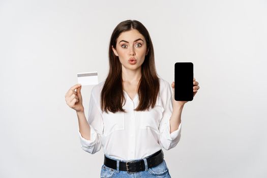 Happy woman showing credit card and smartphone screen, concept of online shopping, buying in app, standing over white background.