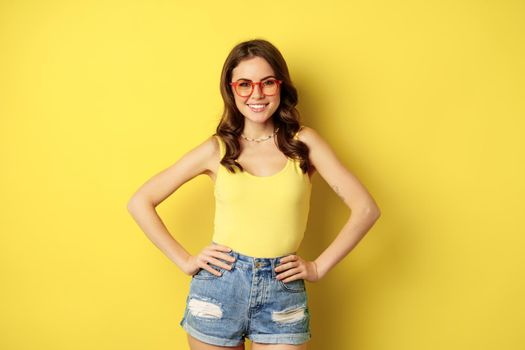 Stylish pretty woman posing in sunglasses and summer clothes, looking happy and carefree, concept of vacation and sunny days, standing over yellow background.