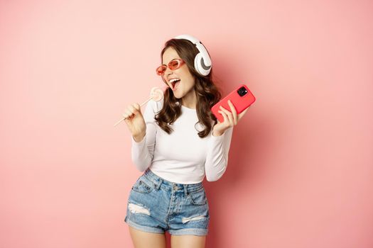 Summer girl laughing, eathing lolipop and listening music in headphones, dancing with smartphone against pink background.