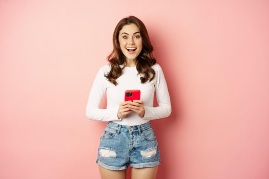 Online shopping and cellphone concept. Excited attractive girl holding mobile phone and smiling, standing over pink background.