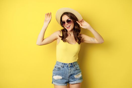 Stylish girl in straw hat and tank top, ready for summer, going on vacation and smiling pleased, standing over yellow background.