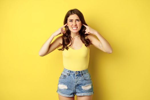 Young stylish woman feeling discomfort from loud noise sound, shut ears with fingers and complaining, standing over yellow background. Copy space