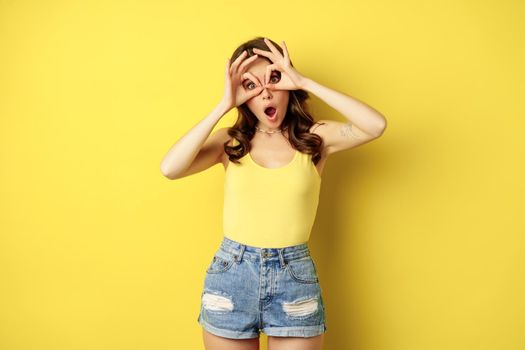 Positive young woman showing okay, ok zero signs, face mask gesture, grimacing and having fun, posing in tank top against yellow background.