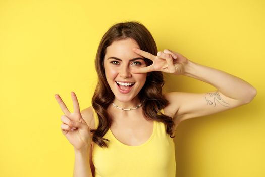 Close up portrait of stylish happy girl, showing peace sign and smiling, posing against yellow background. Copy space