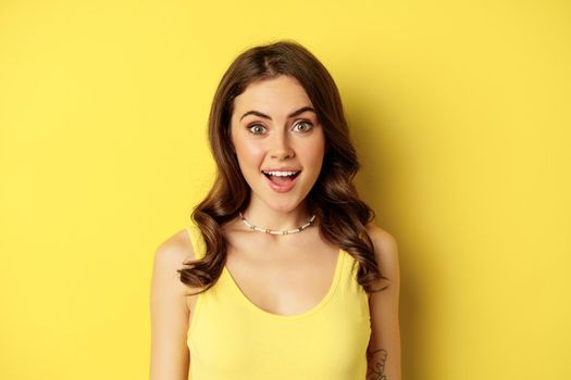 Close up portrait of stylish brunette girl looking amazed, surprised face expression, saying wow, reacting impressed, standing over yellow background.