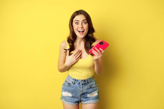 Surprised smiling woman holding mobile phone, reacting amazed and impressed at camera, seeing something awesome, standing against yellow background.