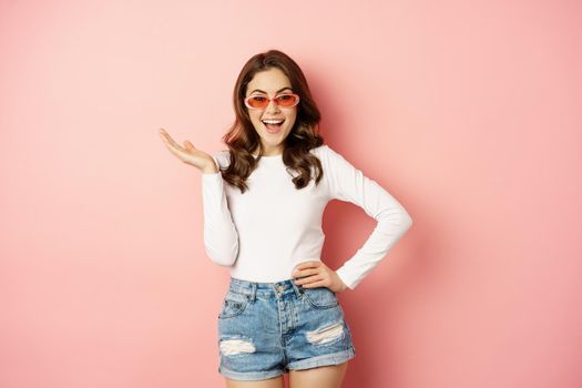 Coquettish and stylish brunette girl, laughing and smiling, wearing sunglasses, wearing white blouse and jeans, pink background. Copy space