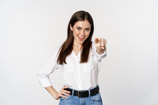 Confident young corporate woman pointing fingers at you camera, inviting, choosing, congratulating, standing over white background.