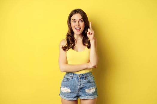 Creative young woman has suggestion, raising finger, eureka sign, has an idea, standing in summer outfit over yellow background. Copy space