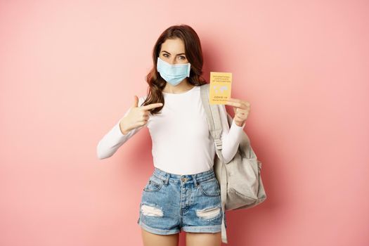 Portrait of woman in medical face mask travelling with covid international vaccination certification, holding backpack, standing over pink background.