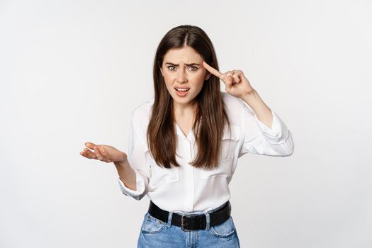 Are you dumb. Annoyed woman scolding, roll finger over head and staring at camera, bothered by someone crazy, standing over white background.