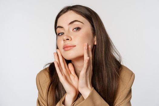 Beauty and skin care. Close up portrait of beautiful woman looking in mirror and touching her clear glowing face, standing over white background.