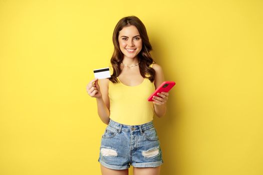 Stylish young brunette woman, holding credit card and smartphone, paying, purchase or order something in store, standing over yellow background.