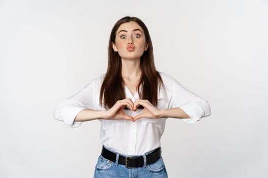 Lovely feminine woman showing heart sign, romantic gesture and smiling with care and tenderness, standing over white background.