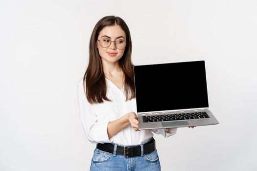 Young woman in glasses showing laptop screen, demonstrating promo on computer, website or store, standing over white background.