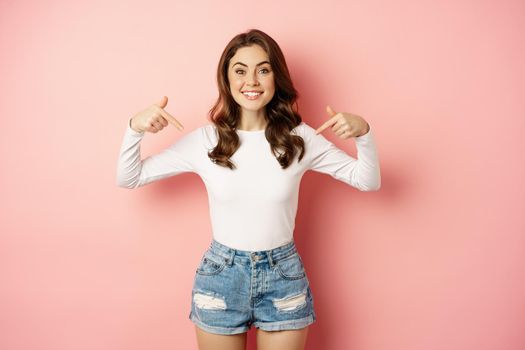 Self-confident young happy woman, stylish girl pointing at herself, smiling satisfied, self-promoting, talking about her, standing against pink background.