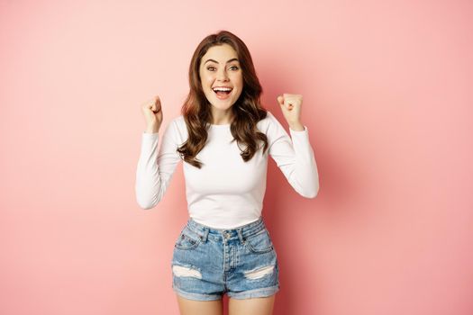 She is a winner. Enthusiastic brunette girl celebrating, jumping from happiness and shouting joyful, achieve goal success, standing over pink background. Victory concept