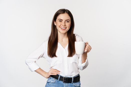 Young moder business woman, office lady holding mug with coffee tea and smiling, standing against white background.