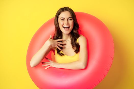 Close up portrait of beautiful brunette woman model inside pink swimming ring, beach accessories and summer vacation concept, yellow background.