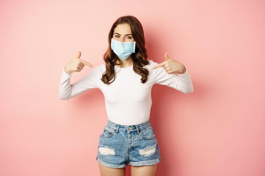 Young woman in medical face mask, pointing fingers at herself, showing logo or banner, smiling at camera, standing over pink background.