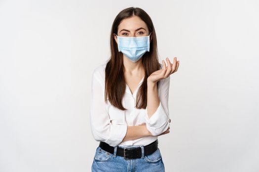 Portrait of beautiful and stylish modern woman, wearing medical face mask during pandemic of covid-19, standing against white background.