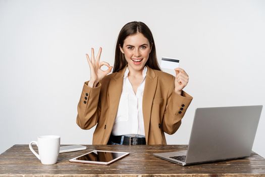Portrait of female entrepreneur sitting at office table, showing credit card and thumbs up, recommending, standing over white background.