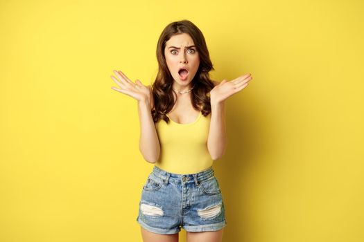 Shocked and frustrated woman gasping, looking overwhelmed and worried, concerned face, standing in summer tank top, yellow background.