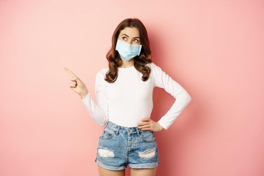 Beautiful stylish woman in medical face mask from covid, pointing finger at upper left corner, showing advertisement, standing over pink background.